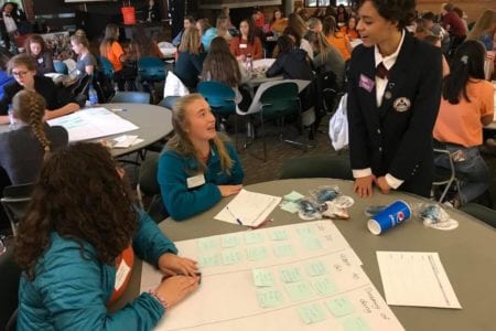 2 HOSA students at a table with sticky notes planning while talking to a state officer in full uniform (other students working at tables in background)
