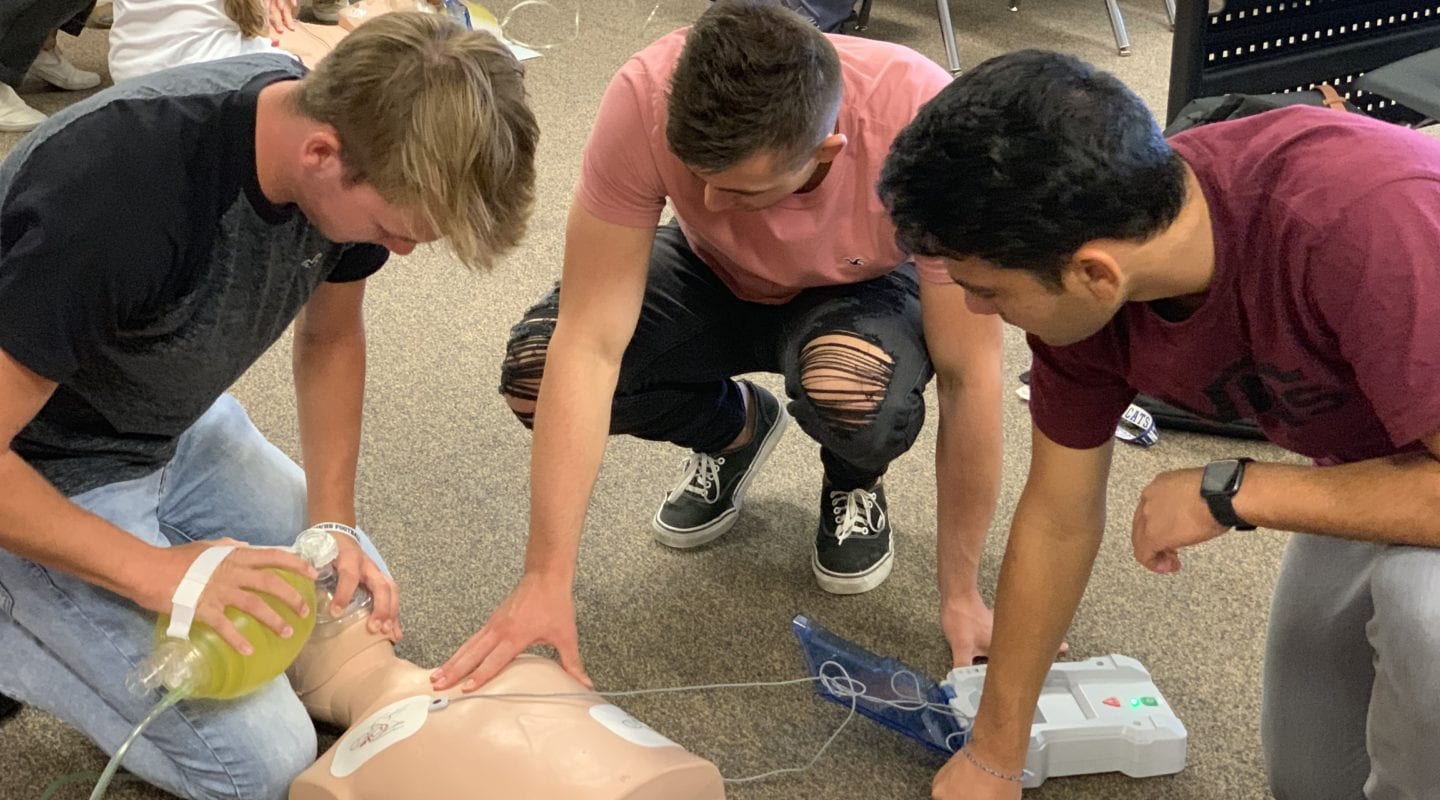 boy using a bag valve mask on a CPR manikin while 2 other boys look on with an AED