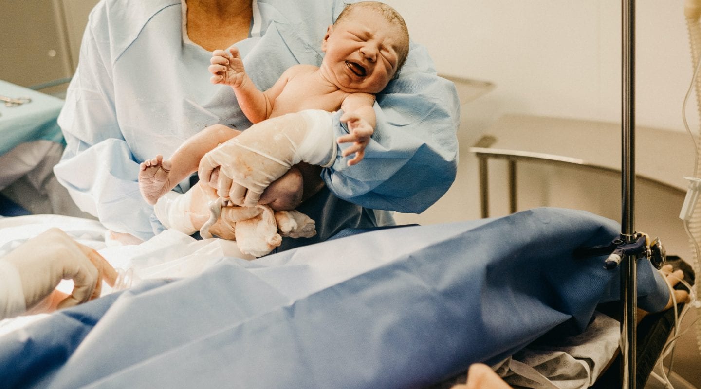 Woman in scrubs holding a newly delivered baby with mom looking on from delivery bed